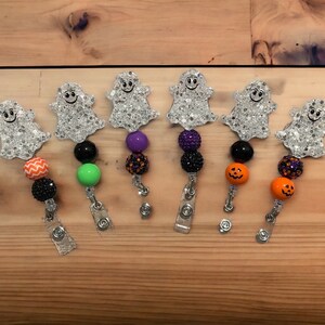 Pu&ograve; includere: Una collezione di porta badge a tema Halloween. Ogni porta badge presenta un ciondolo a forma di fantasma glitterato con un volto sorridente, abbinato a perline colorate in vari design, tra cui zucche arancioni e perline nere. I porta badge sono attaccati a clip argentate.