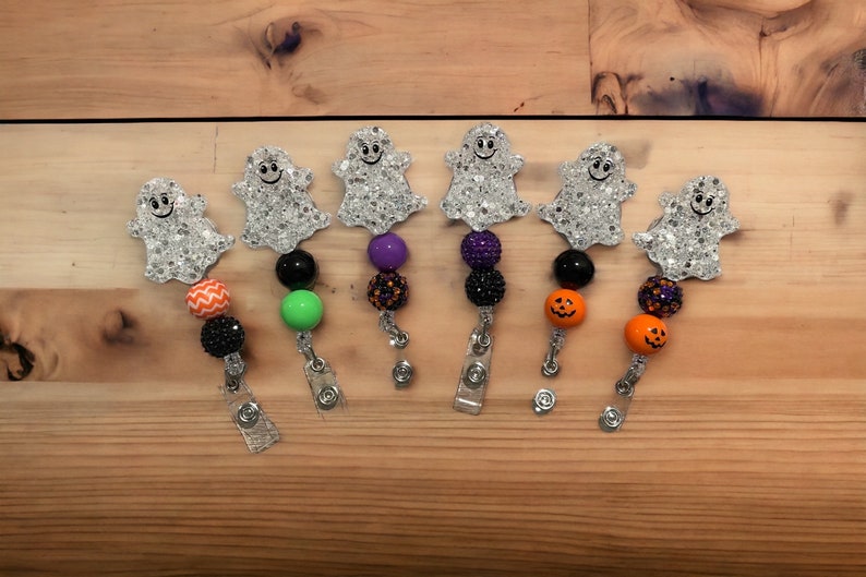 Pu&ograve; includere: Una collezione di porta badge a tema Halloween. Ogni porta badge presenta un ciondolo a forma di fantasma glitterato con un volto sorridente, abbinato a perline colorate in vari design, tra cui zucche arancioni e perline nere. I porta badge sono attaccati a clip argentate.