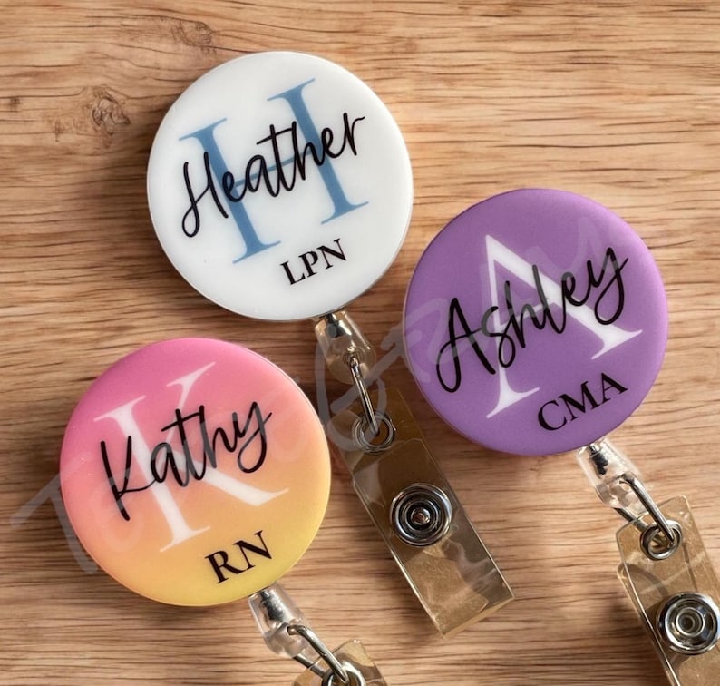 May include: Three round badge holders with retractable cords. The badge holders are decorated with a gradient background and a white initial and name. The first badge holder is white with a blue initial "H" and the name "Heather" with the text "LPN" below. The second badge holder is a pink and orange gradient with a white initial "K" and the name "Kathy" with the text "RN" below. The third badge holder is purple with a white initial "A" and the name "Ashley" with the text "CMA" below.