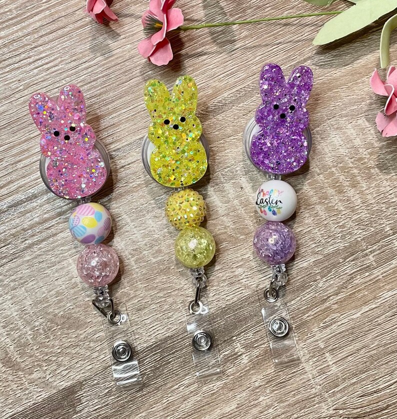 Op de afbeelding: Drie paas-thema badge-rollen met konijnenvormige bedels. De bedels zijn bedekt met glitter en zijn verkrijgbaar in roze, geel en paars. Elke rol heeft een kralenketting met verschillende kleuren en patronen. De paarse rol heeft een witte kraal met de tekst "Prettige Pasen" erop.