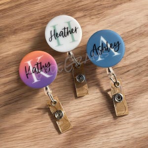 May include: Three personalized badge reels with gold clips. The badge reels are white with green lettering, blue with white lettering, and a gradient of pink and orange with white lettering. The names on the badge reels are Heather, Ashley, and Kathy.
