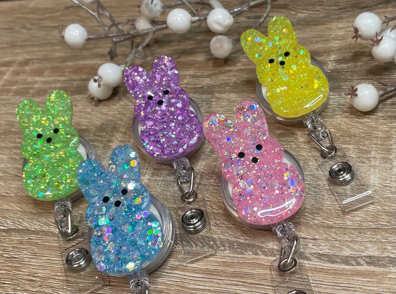 Op de afbeelding: Vijf kleurrijke glitter konijnenvormige intrekbare badgehouders. De houders zijn in tinten groen, paars, roze, blauw en geel. Elke houder heeft een transparante plastic achterkant en een zwart gezicht met twee zwarte stippen voor ogen.