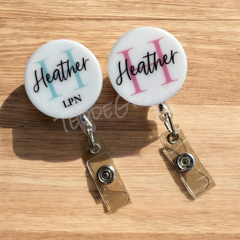 May include: Two white retractable badge holders with a clear plastic strap. The badge holders have a white background with the name "Heather" written in black script with a pink or blue initial "H". One badge holder has the text "LPN" written in black below the name.