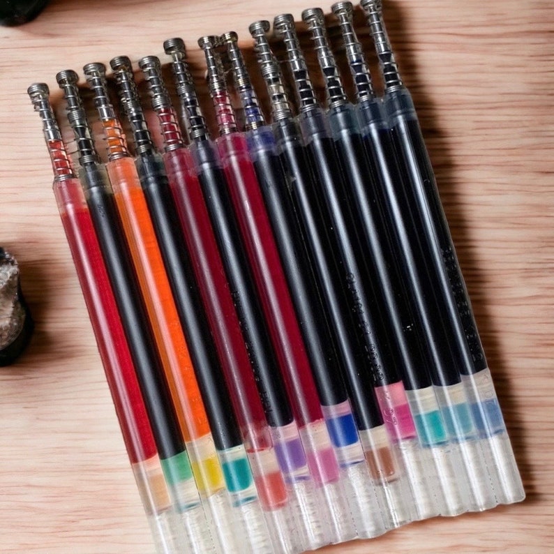 Puede incluir: Primer plano de un conjunto de 17 recambios de tinta de colores para bol&iacute;grafos de gel. Los recambios son negros con puntas de pl&aacute;stico transparente y tienen tinta de diferentes colores en su interior.