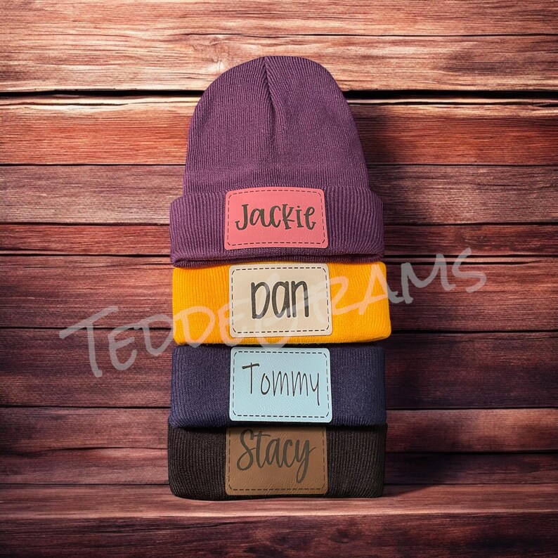 May include: A stack of four beanies with personalized name tags. The beanies are purple, yellow, blue, and brown. The name tags are pink, beige, light blue, and brown. The names on the tags are Jackie, Dan, Tommy, and Stacy.