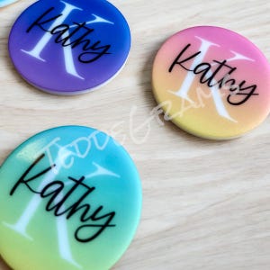 May include: Three round phone grips with a gradient background. Each grip has the name "Kathy" written in black cursive with a white letter "K". The grips are purple, yellow, and green.