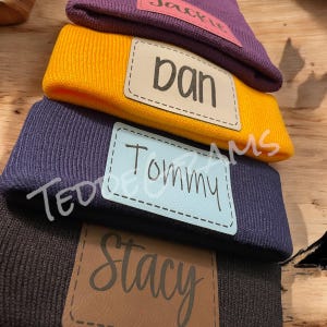 May include: Stack of four knitted beanies with personalized leather patches. The patches are brown, beige, blue, and pink. The names on the patches are Stacy, Tommy, Dan, and Jackie.