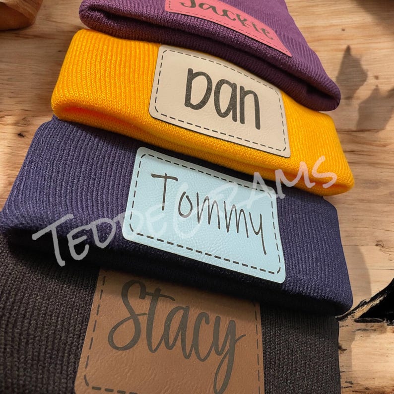 May include: Stack of four knitted beanies with personalized leather patches. The patches are brown, beige, blue, and pink. The names on the patches are Stacy, Tommy, Dan, and Jackie.