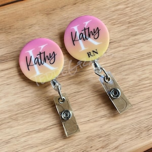 May include: Two pink and yellow gradient retractable badge holders with a white letter "K" and the name "Kathy" in black script. The bottom badge holder also has the letters "RN" in black.