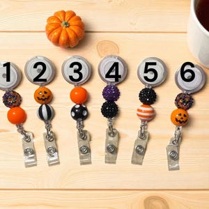 Pu&ograve; includere: Sei porta badge a tema Halloween con perline e ciondoli decorativi. Ogni porta badge ha un design diverso, tra cui una zucca, una lanterna e perline a righe. Una piccola zucca arancione &egrave; sopra.