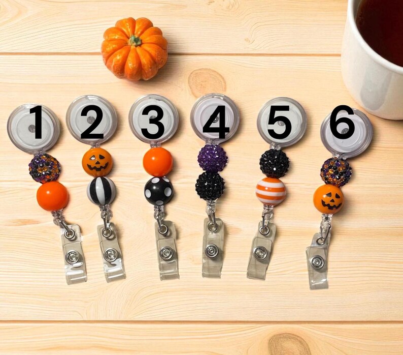 Pu&ograve; includere: Sei porta badge a tema Halloween con perline e ciondoli decorativi. Ogni porta badge ha un design diverso, tra cui una zucca, una lanterna e perline a righe. Una piccola zucca arancione &egrave; sopra.