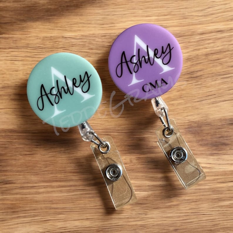 May include: Two round, personalized badge holders with retractable reels. One is light green with the name "Ashley" and the other is purple with the name "Ashley" and the letters "CMA".