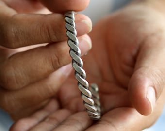 May include: A silver-colored, braided cuff bracelet. The bracelet has a twisted rope design. The open cuff design allows for easy wear. The bracelet is held in open hands.