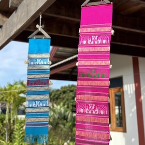 May include: Two colorful, rectangular textile wall hangings. One is bright blue with gold, red, and white patterns. The other is hot pink with gold and green patterns. Both have fringe and are suspended from wooden structures.