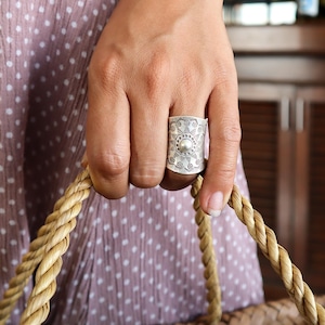 May include: A wide, silver ring with an embossed design and a central pearl-like bead. The ring is worn on a finger, and the hand is holding a woven basket. The ring's design includes floral and geometric patterns.
