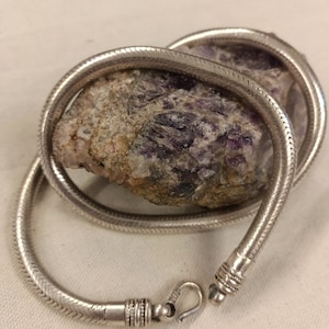 Pu&ograve; includere: Una collana a catena di serpente d'argento con una chiusura. La catena &egrave; avvolta attorno a una pietra viola e grigia.