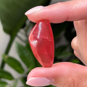 May include: A smooth, polished, pink and red gemstone with a pointed tip. The gemstone is held between two fingers.
