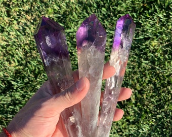 May include: Three amethyst crystal points with a purple and white color gradient. The points are held in a hand.