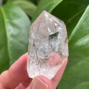 May include: A clear quartz crystal with black and gold inclusions. The crystal is faceted and has a pointed tip.