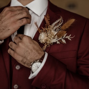 May include: A man in a burgundy suit adjusts his black tie. He is wearing a white shirt and a watch. A dried flower boutonniere is pinned to his lapel.