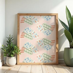 May include: A framed print featuring a repeating pattern of stylized floral and geometric shapes in shades of blue, green, red, and cream on a light pink background. The wooden frame adds a natural touch to the artwork. The print is displayed on a wooden floor with potted plants.