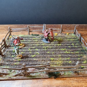 May include: A miniature tabletop gaming scene with three painted figures in a field surrounded by a wooden fence. The figures are a warrior with a sword, a knight with a shield, and a smaller figure with a staff. The field is painted with brown and green colors to represent dirt and grass.