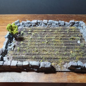May include: A miniature stone wall surrounds a small field of green plants. The field is made of a brown material with a textured surface. A small green bush is growing in the corner of the field.