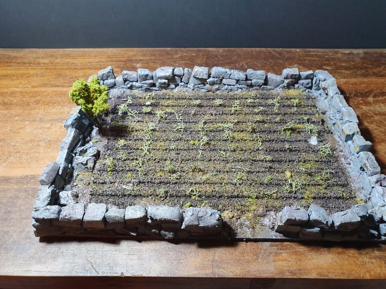 May include: A miniature stone wall surrounds a small field of green plants. The field is made of a brown material with a textured surface. A small green bush is growing in the corner of the field.