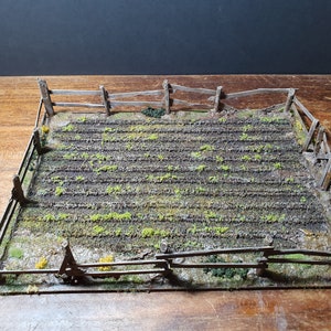 May include: A miniature model of a fenced-in field with rows of green plants growing in the dirt. The field is surrounded by a wooden fence with a rustic, weathered look.