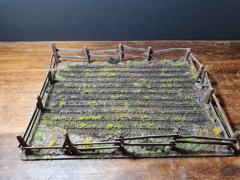 May include: A miniature model of a fenced-in field with rows of green plants growing in the dirt. The field is surrounded by a wooden fence with a rustic, weathered look.