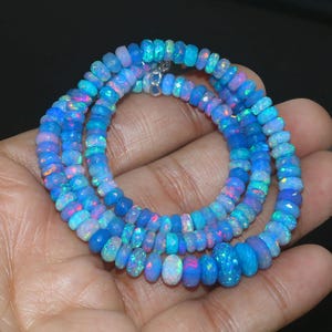 Op de afbeelding: Een ketting van gefacetteerde, ovale opalen in tinten blauw, roze en wit. De kralen zijn geregen met een zilveren sluiting. De ketting wordt gepresenteerd tegen een donkere achtergrond.