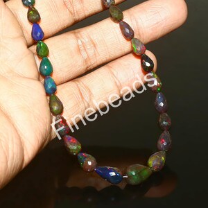 May include: A strand of faceted, teardrop-shaped opal beads. The opals display a variety of colors, including blue, green, red, and black. The beads are strung together, creating a necklace or bracelet.