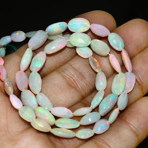 Puede incluir: Un collar de cuentas de ópalo ovaladas. Los ópalos muestran un color base blanco lechoso con destellos de colores iridiscentes, incluyendo rosa, verde y azul. Las cuentas están ensartadas, mostrando sus superficies pulidas y variaciones naturales.