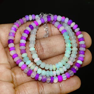 Puede incluir: Collar de varias hebras con pequeñas cuentas redondas en tonos morado, blanco y azul claro. El collar tiene un cierre plateado. Las cuentas están ensartadas para crear un efecto de capas.