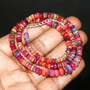 May include: A vibrant necklace composed of small, faceted opal beads in shades of red, orange, purple, and green. The beads are strung together with a silver chain clasp. The necklace is displayed on a hand against a dark background.