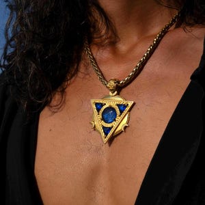 May include: A gold-colored pendant necklace with a triangular design. The pendant features a central blue stone and is surrounded by blue accents. The chain is a thick, braided style. The necklace is worn against a dark background.