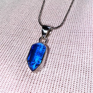 Könnte beinhalten: Eine silberne Halskette mit einem Anhänger, der einen facettierten, rechteckigen, leuchtend blauen Edelstein zeigt. Der Anhänger ist in eine silberne Lünette gefasst und hängt an einer zarten Silberkette. Der Hintergrund ist ein hellrosa, strukturiertes Gewebe.