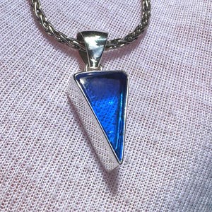 Può includere: Ciondolo in argento con un inserto triangolare blu intenso, sospeso a una catena d'argento. Il design geometrico e il colore vibrante del ciondolo creano un contrasto sorprendente. La catena ha una trama intrecciata.