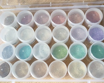 May include: A clear plastic storage box filled with 24 small containers of various colored glitter. The containers are arranged in a grid pattern.