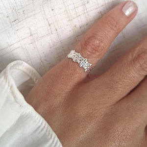 Könnte beinhalten: Ein silberner Ring mit einem durchgehenden Band aus kleinen, geprägten Blumenformen. Das Metall hat eine polierte, reflektierende Oberfläche und das florale Muster verläuft vollständig um den Ring herum, was ein strukturiertes Design ergibt.