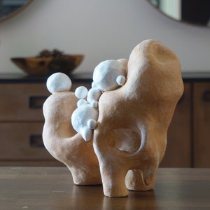 May include: Abstract ceramic sculpture in shades of tan and white. The artwork features two figures embracing, with smaller white spheres clustered between them. The sculpture is displayed on a dark wooden surface.