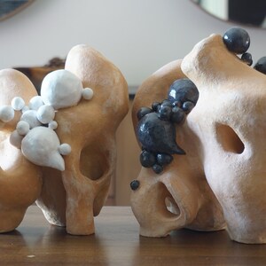 May include: Two abstract sculptures with organic forms. The sculptures are a light tan colour, with white and dark grey spherical accents. The sculptures have holes and rounded shapes, and are displayed on a wooden table.