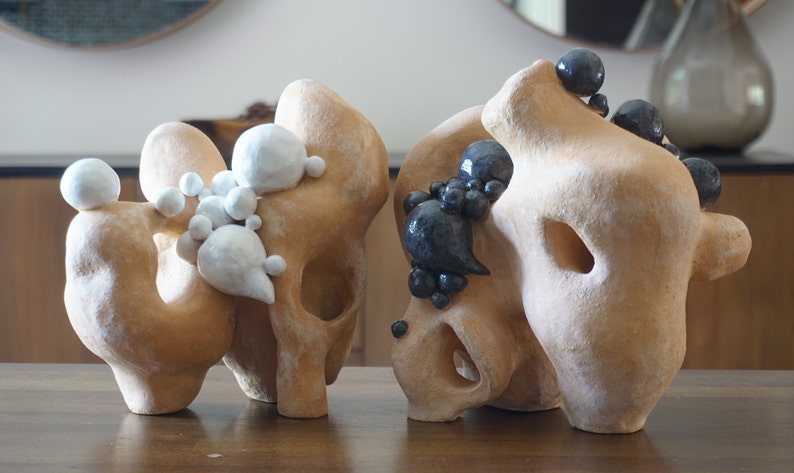 May include: Two abstract sculptures with organic forms. The sculptures are a light tan colour, with white and dark grey spherical accents. The sculptures have holes and rounded shapes, and are displayed on a wooden table.