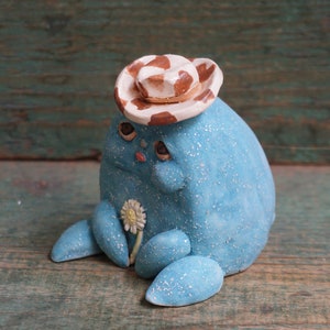 May include: A blue ceramic figurine wearing a white and brown cowboy hat, holding a white flower. The figurine has a sparkly finish and a whimsical expression.