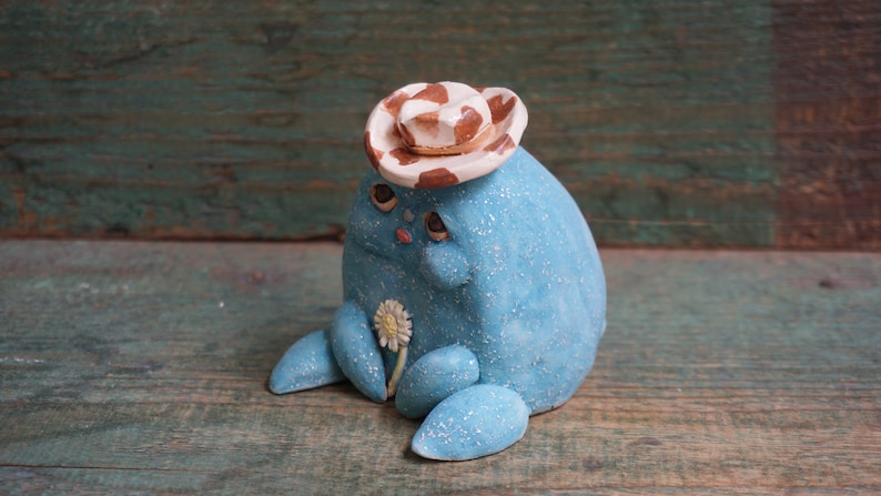 May include: A blue ceramic figurine wearing a white and brown cowboy hat, holding a white flower. The figurine has a sparkly finish and a whimsical expression.