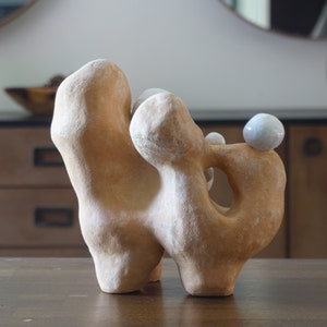 May include: Abstract ceramic sculpture in a light brown colour with a textured surface. The sculpture has a unique shape with rounded forms and a hole. Two small white spheres are placed on top.