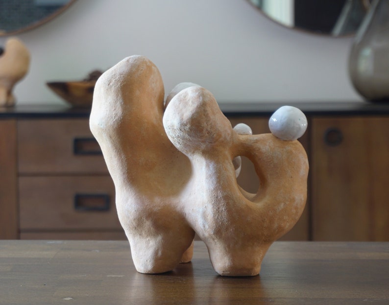 May include: Abstract ceramic sculpture in a light brown colour with a textured surface. The sculpture has a unique shape with rounded forms and a hole. Two small white spheres are placed on top.