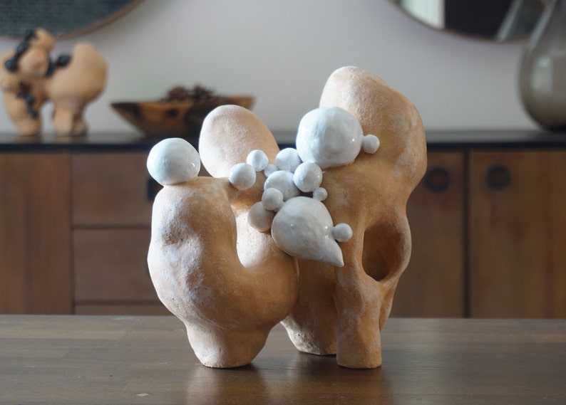 May include: Abstract ceramic sculpture in tan with white, bulbous accents. The organic form features rounded shapes and a textured surface. A second, similar sculpture is visible in the background, along with a wooden bowl and a cabinet.