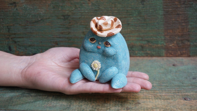 May include: A small, blue, ceramic figurine of a creature wearing a brown and white cowboy hat and holding a white flower. The figurine is sitting on a person's hand.