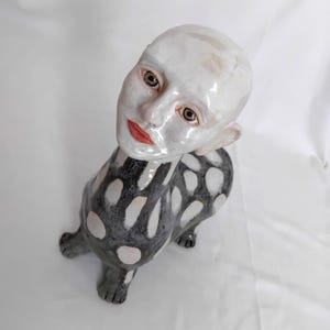 May include: A ceramic sculpture featuring a human-like head with a glossy white finish, brown eyes, and red lips, attached to a four-legged animal body. The body is decorated with a dark gray pattern of irregular white spots across its surface.